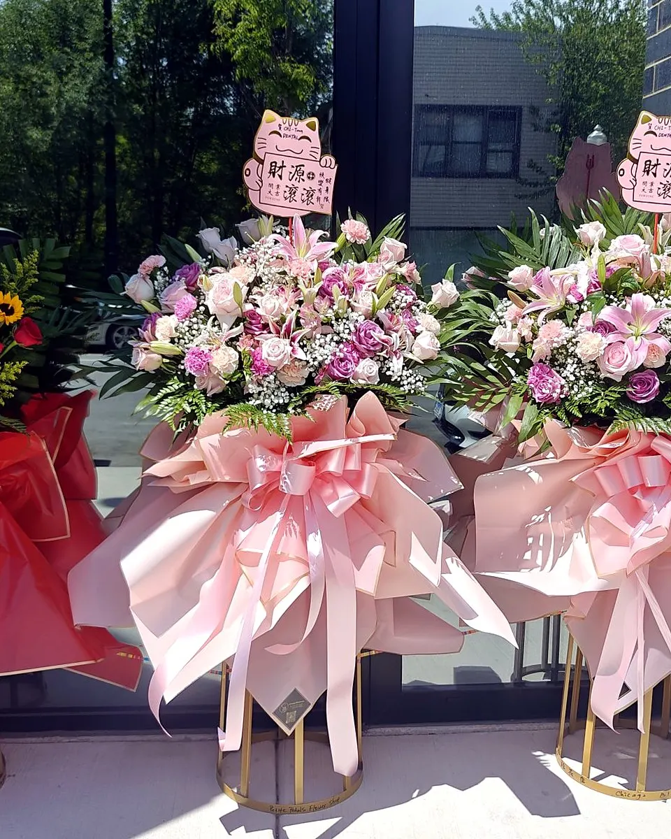 Three grand opening flower stands with pink, red, and yellow flowers outside a storefront
